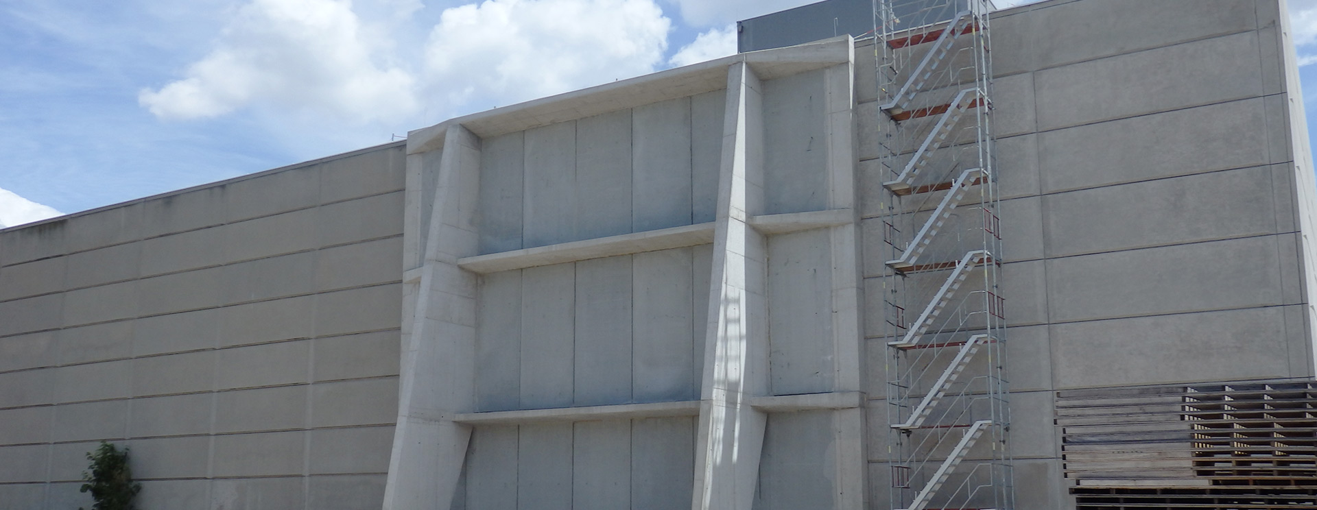 Großes graues Gebäude aus Beton mit einer äußeren Metalltreppe und einem Stapel Holzpaletten daneben, unter einem bewölkten Himmel.
