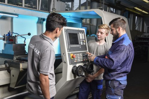 Das Bild zeigt 3 Lehrlinge in Arbeitskleidung, die in einer Werkstatt eine Maschine inspizieren oder reparieren.