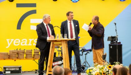 Drei Männer in Anzügen stehen auf einer Bühne vor einer gelben Wand mit einem Mikrofon, umgeben von Blumen und Instrumenten.